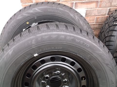 1999 Mercury Grand Marquis - Four Bridgestone Blizzak WS70 tires mounted on black steel wheels are stacked against a brick wall.