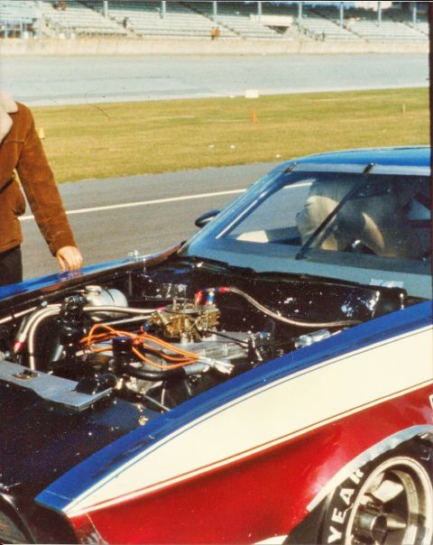 1973 Ford Mustang - The engine bay of a 1973 Ford Mustang, a prominent carburetor and racing stripes, is shown at a track.
