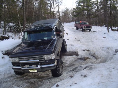 1993 Ford Bronco - Two SUVs navigate a challenging snowy and rocky path in a wooded area.