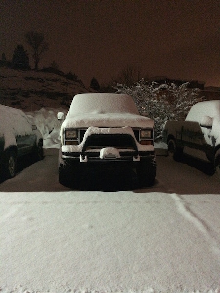 1981 Ford Bronco - Snowfall covers a Ford Bronco and surrounding vehicles parked outside at night.