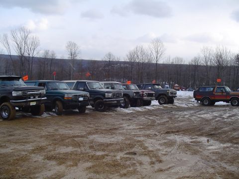 1993 Ford Bronco - Several Ford Broncos and F-Series trucks are parked in a muddy, outdoor lot under a cloudy sky.