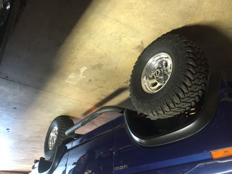 1982 Ford Bronco - The side of a blue SUV is shown with large off-road tires and chrome wheels, parked inside a garage.