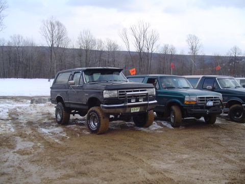 1993 Ford Bronco - A lifted 1993 Ford Bronco is parked in a muddy area alongside other vehicles, with trees and snow in the background.