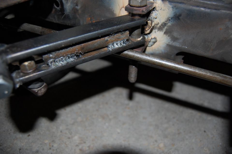 Close-up of a metal frame with visible welds and bolts. A thin rod runs through the frame