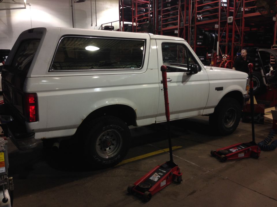 White Ford Bronco side view in a tire shop with two red floor jacks and tire racks behind