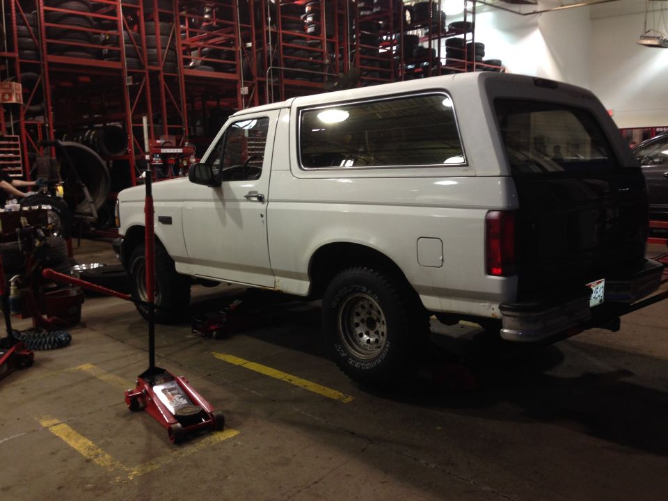 White Ford Bronco on jacks inside a tire shop, rear three-quarter view with red tire racks behind