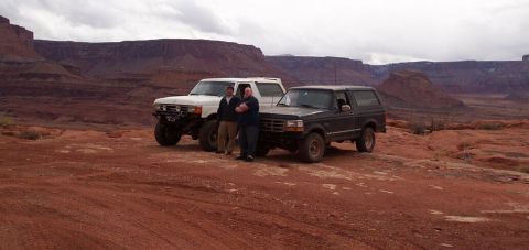1990 Ford Bronco - Two off-road vehicles are parked in a desert landscape with red rock formations under a cloudy sky.