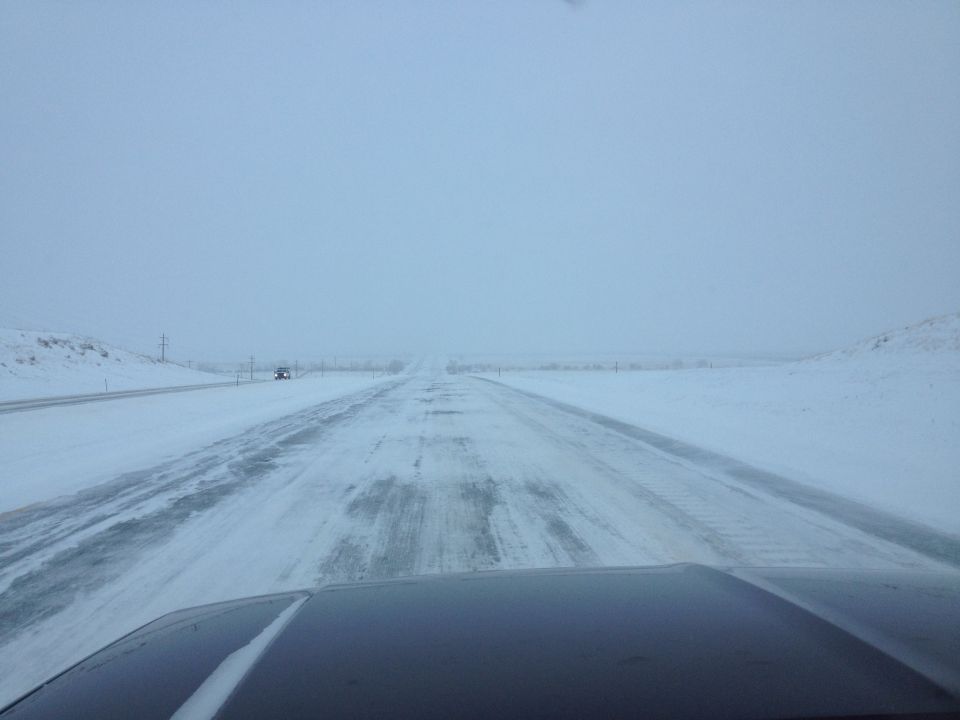 Driving on a snow-covered highway with another vehicle in the distance. The foreground shows the hood of a dark vehicle