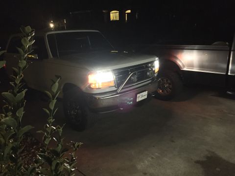 1995 Ford Bronco - A white Ford Bronco is parked next to another truck at night, with its headlights on.