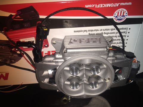1982 Ford Bronco - A close-up, low-angle view of an MSD throttle body with a black braided wire harness connected. The throttle body is silver and has four vi…