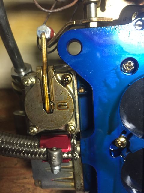 1982 Ford Bronco - A close-up view of a blue-coated metal part attached to a carburetor, with a braided fuel line and a red fitting visible below.