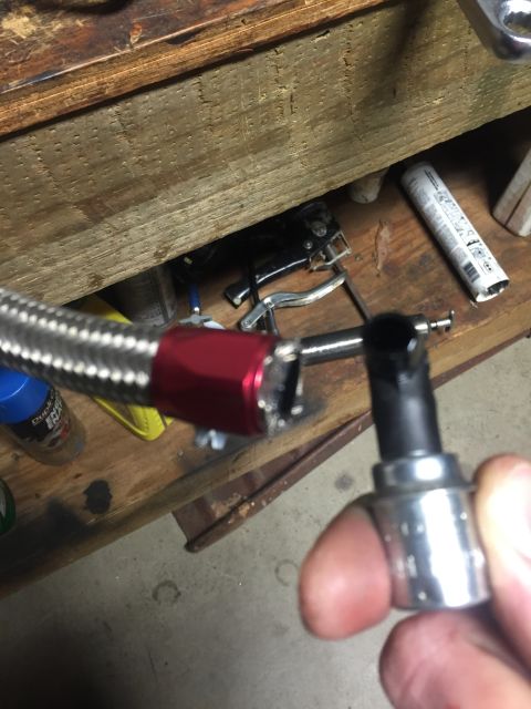 1982 Ford Bronco - A close-up view of a braided metal hose with a red fitting, held by a hand with a socket wrench. The hose appears to be a vehicle part, pos…