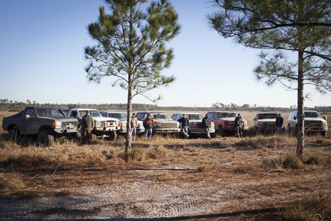 1992 Ford Bronco - A group of people stand around a line of pickup trucks and SUVs parked in a dry, grassy field under a clear blue sky.