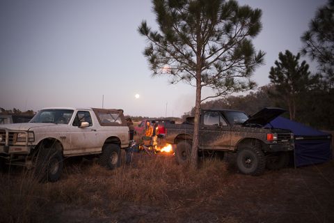 1992 Ford Bronco - Two rugged, mud-splattered pickup trucks and a Bronco are parked in a grassy field at dusk, with a campfire burning between them. A full mo…