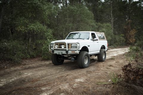 1992 Ford Bronco - A lifted white Ford Bronco is driving on a dirt road surrounded by trees. The vehicle has large tires and a brush guard on the front.