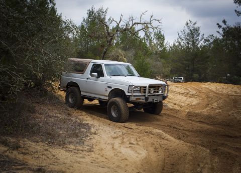 1992 Ford Bronco - A white 1992 Ford Bronco with a tan soft top is parked on a sandy track surrounded by trees. The vehicle has large off-road tires and a bru…