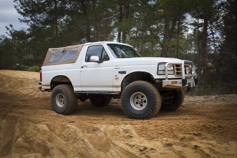 1992 Ford Bronco - A lifted white 1992 Ford Bronco with a tan soft top drives on a sandy track surrounded by trees. The vehicle has large off-road tires and a…