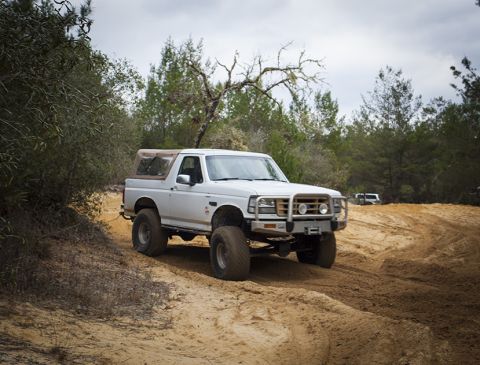 1992 Ford Bronco - A white 1992 Ford Bronco with a brown soft top is driving on a sandy trail through a wooded area. The vehicle has large off-road tires and…