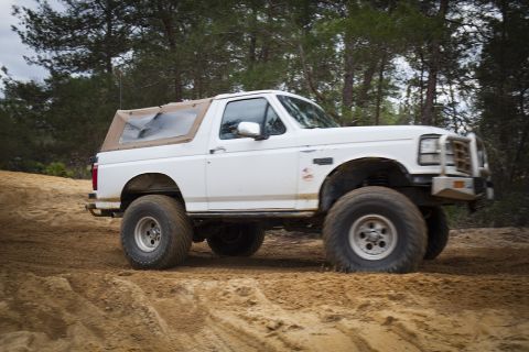 1992 Ford Bronco - A white Ford Bronco with a tan soft top is driving on a sandy trail surrounded by pine trees. The vehicle has large off-road tires and a br…