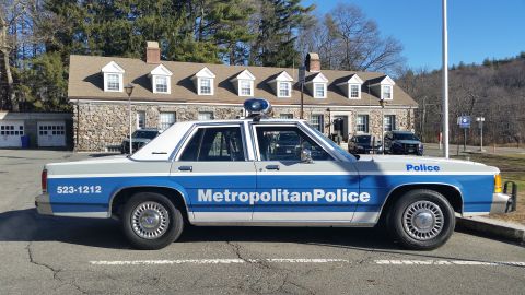 1991 Ford LTD Crown Victoria - Photo of a vintage Metropolitan Police Ford LTD Crown Victoria cruiser parked beside a stone police station.