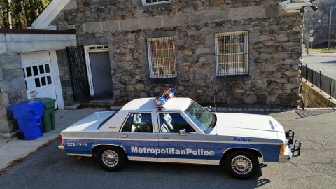 1991 Ford LTD Crown Victoria - Photo of a vintage Ford Metropolitan Police patrol car parked in front of a stone garage building, not a technical document.