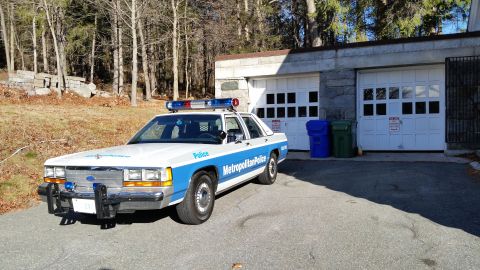 1991 Ford LTD Crown Victoria - Photo of a vintage Ford Metropolitan Police patrol car with front push bar parked by garages, not a technical document.
