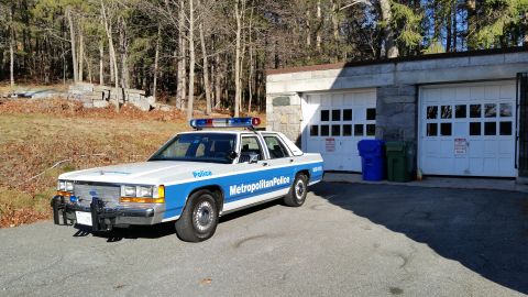 1991 Ford LTD Crown Victoria - Photo of a vintage Ford Metropolitan Police patrol car parked near garage doors in a wooded lot, not a technical document.