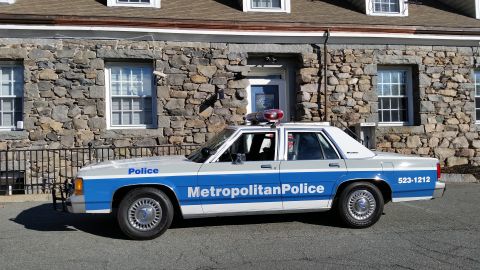 1991 Ford LTD Crown Victoria - Photo of a vintage Ford Metropolitan Police patrol car shown in side profile by a stone building, not a technical document.