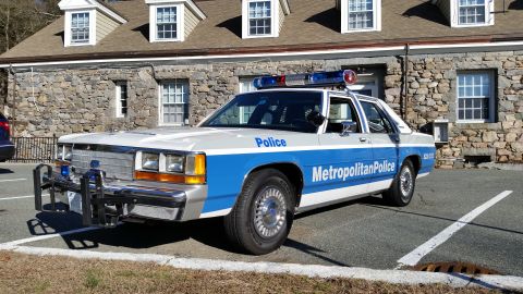 1991 Ford LTD Crown Victoria - Photo of a vintage Ford Metropolitan Police patrol car with front push bar by a stone building, not a technical document.