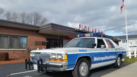 1991 Ford LTD Crown Victoria - Photo of a vintage Ford Metropolitan Police patrol car parked outside Florian Hall under a cloudy sky, not a technical document.