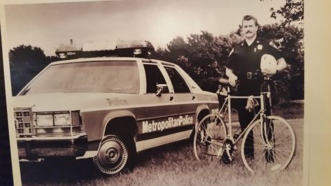 1991 Ford LTD Crown Victoria - Sepia photo of a Metropolitan Police bicycle officer standing beside a Crown Victoria cruiser, not a technical document.