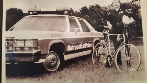 1991 Ford LTD Crown Victoria - Sepia photo of a Metropolitan Police bicycle officer with a Crown Victoria cruiser in a field, not a technical document.