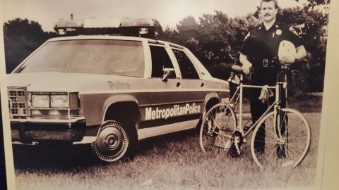 1991 Ford LTD Crown Victoria - Sepia photo of a Metropolitan Police bicycle officer standing by a Crown Victoria cruiser, not a technical document.