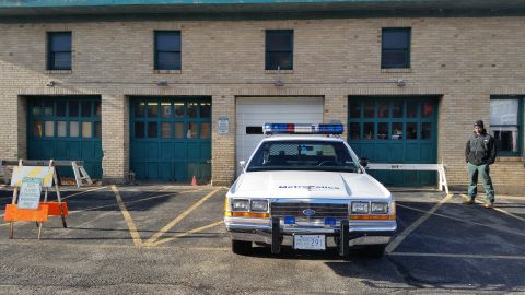 1991 Ford LTD Crown Victoria - Photo of a vintage Metropolitan Police Ford Crown Victoria parked in front of a brick station with green garage doors.