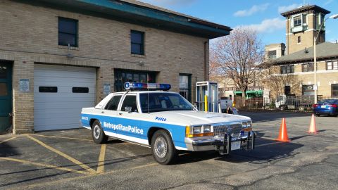 1991 Ford LTD Crown Victoria - Photo of a blue and white Metropolitan Police Crown Victoria parked beside a historic brick station.