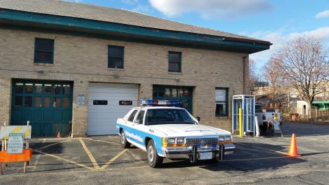 1991 Ford LTD Crown Victoria - A white car is parked in front of a brick building with garage doors.