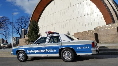 1991 Ford LTD Crown Victoria - A 1991 Ford Crown Victoria police car is seen from the side, parked near a large building with an arched structure.