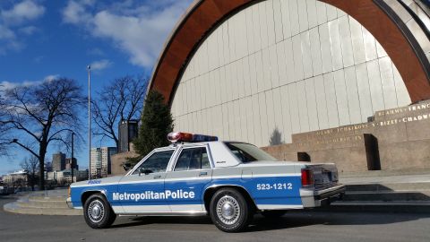 1991 Ford LTD Crown Victoria - A 1991 Ford Crown Victoria police car is parked in front of a modern building with a large arched facade.