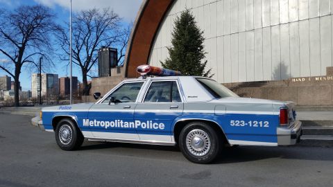 1991 Ford LTD Crown Victoria - A police car with a light bar on the roof is parked on a street with buildings in the background.