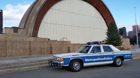 1991 Ford LTD Crown Victoria - A 1991 Ford Crown Victoria police car is parked in front of a modern building with a large arched facade.