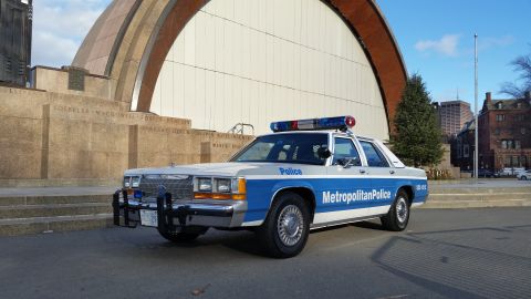 1991 Ford LTD Crown Victoria - A police car is parked in front of a large building with a curved facade and inscribed stone walls.