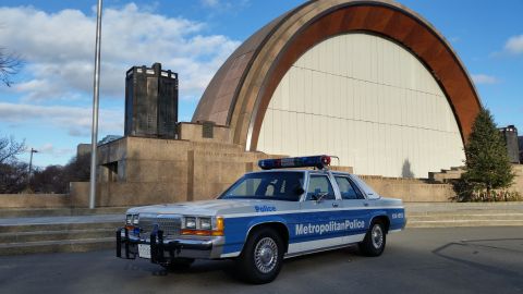 1991 Ford LTD Crown Victoria - A 1991 Ford Crown Victoria police car is parked in front of a distinctive modern building with a large arched facade.