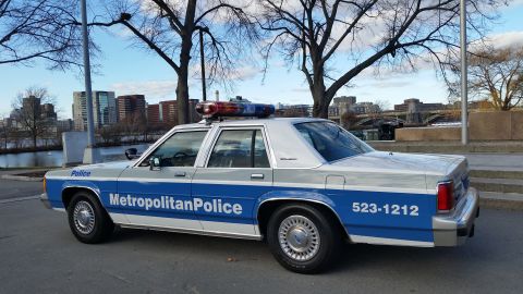 1991 Ford LTD Crown Victoria - A police car with a light bar on the roof is parked near a body of water with a city skyline in the background.