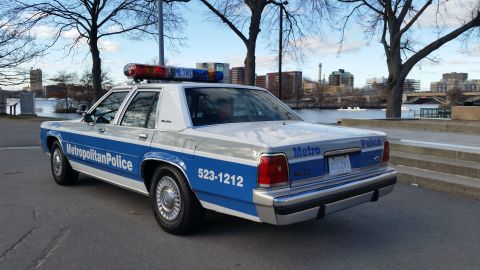 1991 Ford LTD Crown Victoria - A police car with 'Metropolitan Police' and '523-1212' markings is parked near a body of water with a city skyline in the background.