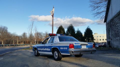 1991 Ford LTD Crown Victoria - A police car with 'Metropolitan Police' markings is parked near a building under a partly cloudy sky.