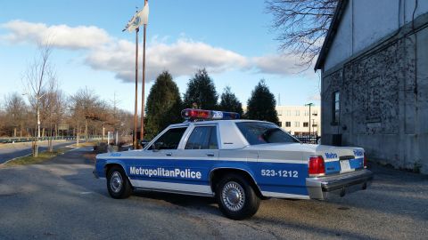 1991 Ford LTD Crown Victoria - A 1991 Ford Crown Victoria police car is seen from the rear quarter, parked on an asphalt surface with a building and trees in the backgrou…