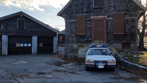 1991 Ford LTD Crown Victoria - A police car sits parked in front of an abandoned building with a garage to its left.
