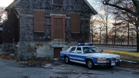 1991 Ford LTD Crown Victoria - A police car is parked next to an old, abandoned building with boarded-up windows.