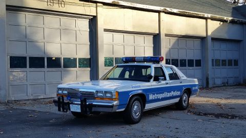 1991 Ford LTD Crown Victoria - Metropolitan Police car, likely a 1991 Ford Crown Victoria, parked outside a building with garage doors.