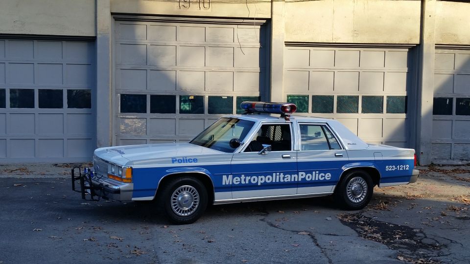 1991 Ford Crown Victoria police car, white and blue, with light bar, parked in front of garage doors